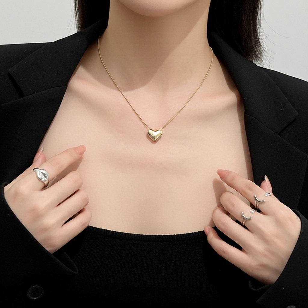 Gold heart-shaped necklace worn by a person with black blazer and rings on fingers.